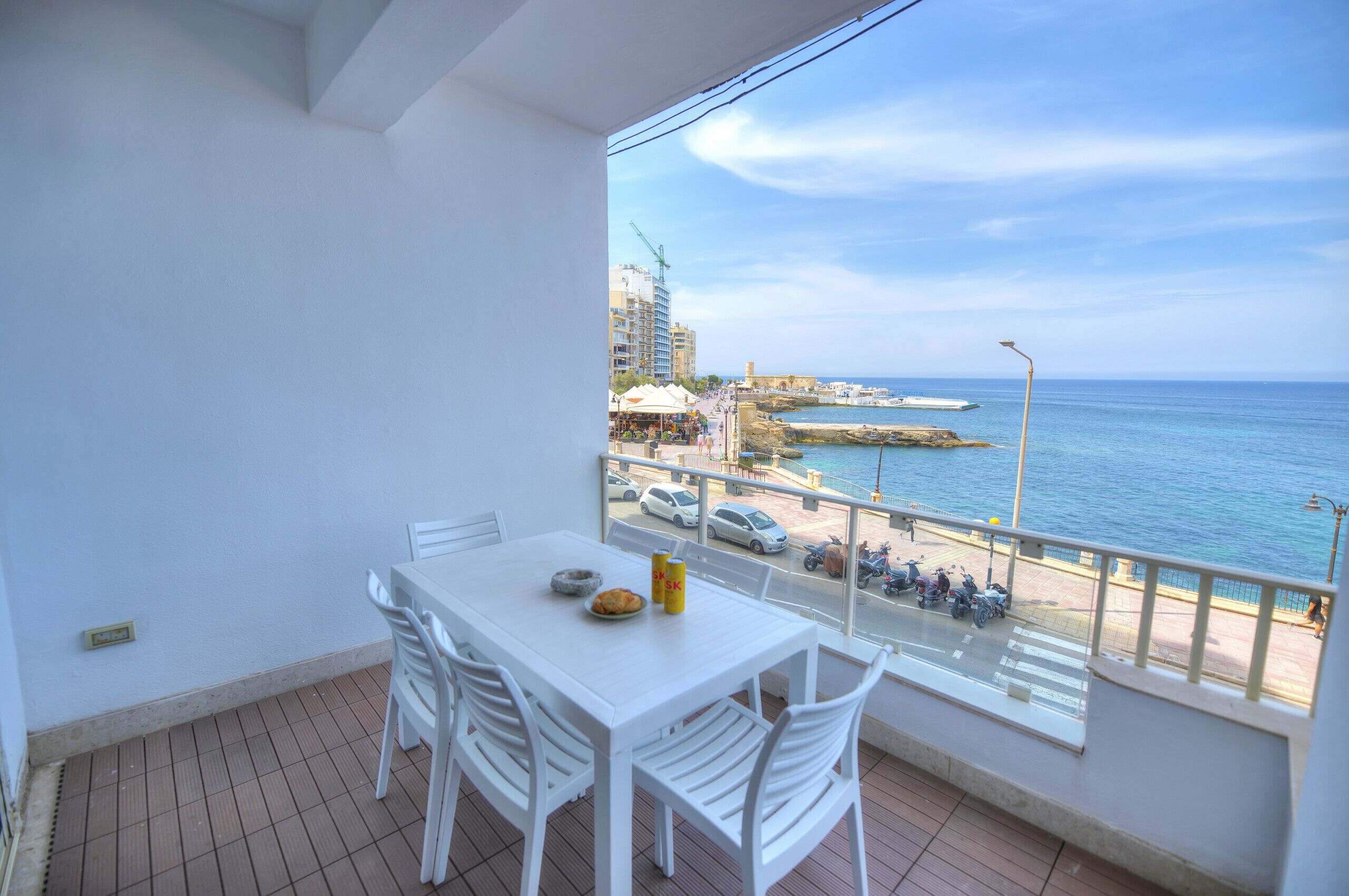 Un balcón con mesa y sillas blancas, con vista a una zona costera que incluye edificios, un muelle y coches aparcados.