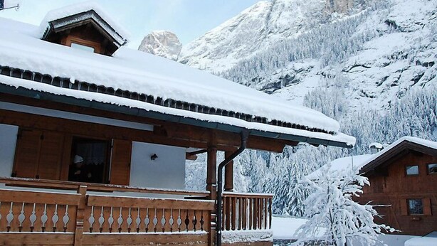 Una cabaña de madera con balcón, rodeada de montañas y árboles cubiertos de nieve.