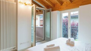 A bedroom with a wooden ceiling, a radiator, a bed with white linens and towels, and a sliding glass door leading to an outdoor balcony with a view of trees. - Luxury House by Interhome (Colico)