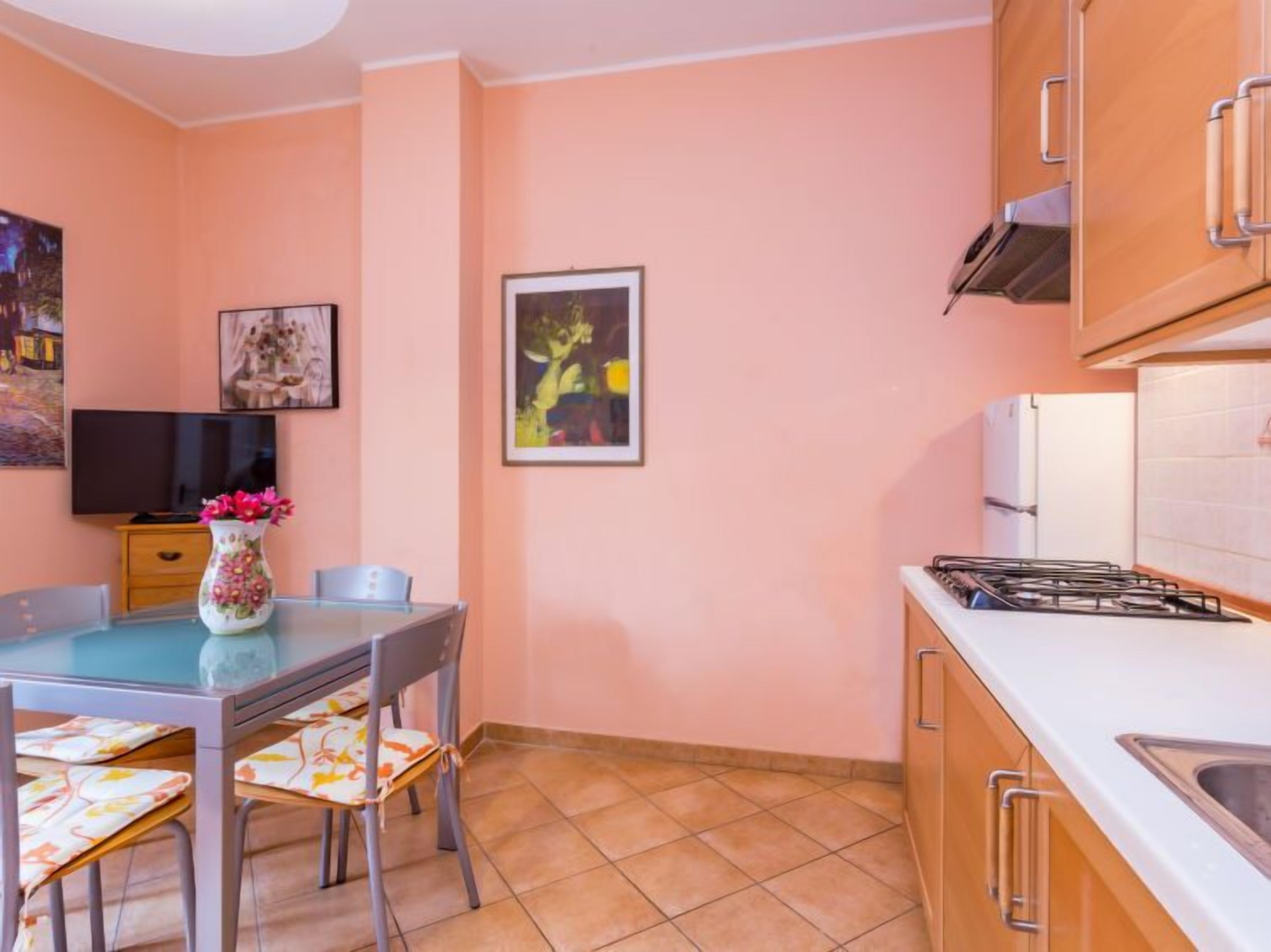 A compact kitchen with a gas stove, a sink, and wooden cabinets. There is a dining table with chairs and a vase with flowers.