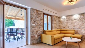 Un salón moderno con pared de piedra, un sofá amarillo, una mesa redonda blanca y un ventanal de vidrio que da a un espacio al aire libre con mesa y sillas.