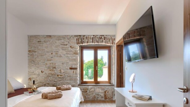 Un dormitorio con pared de piedra, una cama con colcha blanca, una mesita de noche con un libro y un espejo, y un televisor montado en la pared.