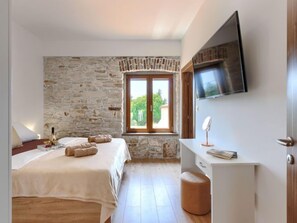 Ein Schlafzimmer mit Steinwand, einem Bett mit weißer Bettdecke, einem Nachttisch mit Buch und Spiegel sowie einem an der Wand befestigten Fernseher.
