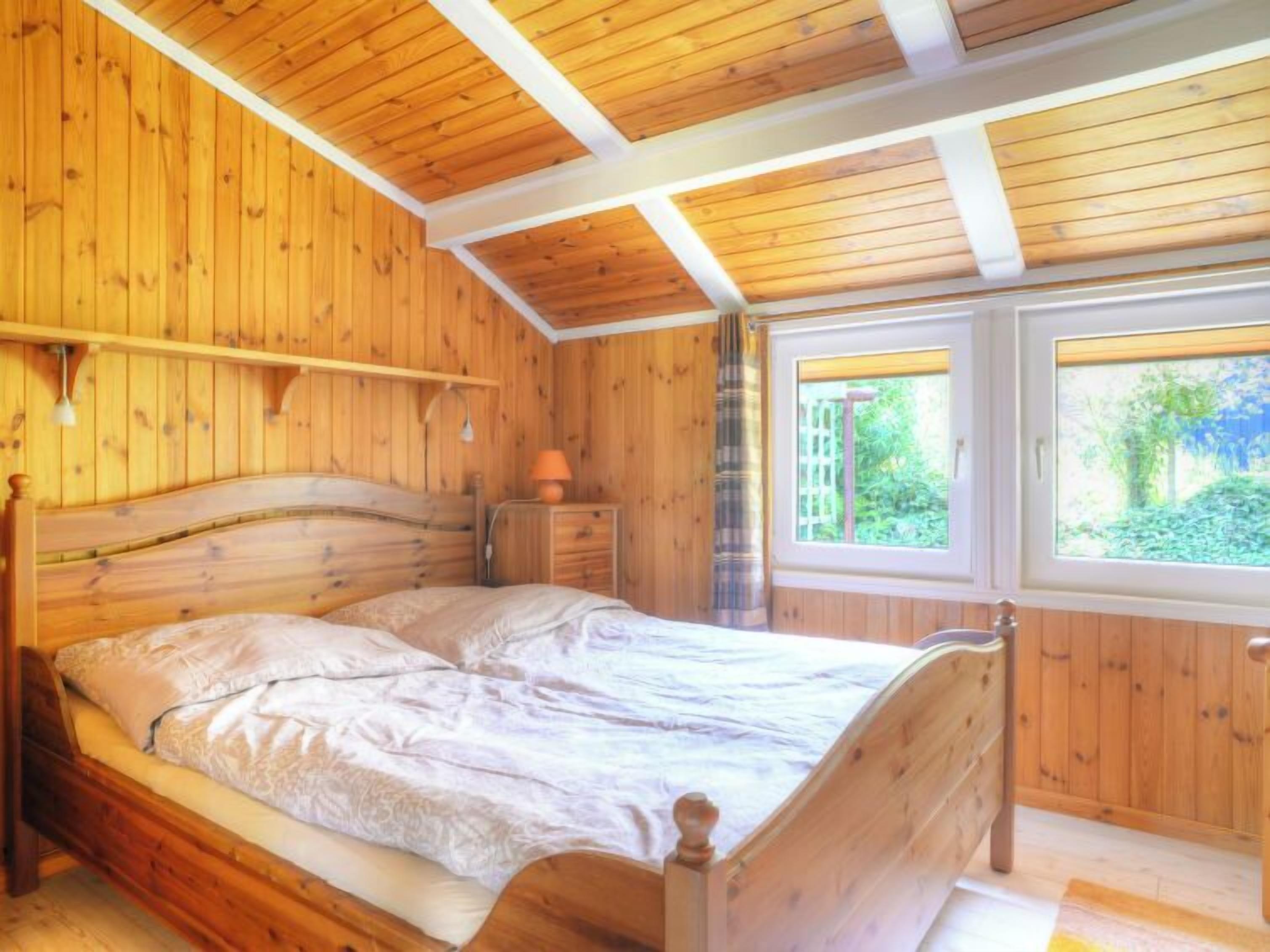 Een slaapkamer met houten lambrisering, een bed, een nachtkastje, een lamp en een raam met uitzicht op groen.