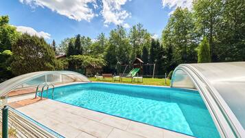 A swimming pool with a slide, surrounded by a covered deck and a fenced area with chairs.