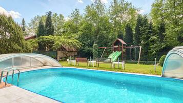 A swimming pool with a slide, surrounded by a fenced area with benches and a covered seating area.