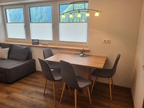 A modern living room with a wooden dining table, four grey chairs, a grey sofa, and a window with blinds.