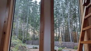 Habitación con un ventanal grande que ofrece vistas a un bosque. Se observa una escalera de madera y una silla de mimbre con cojines.