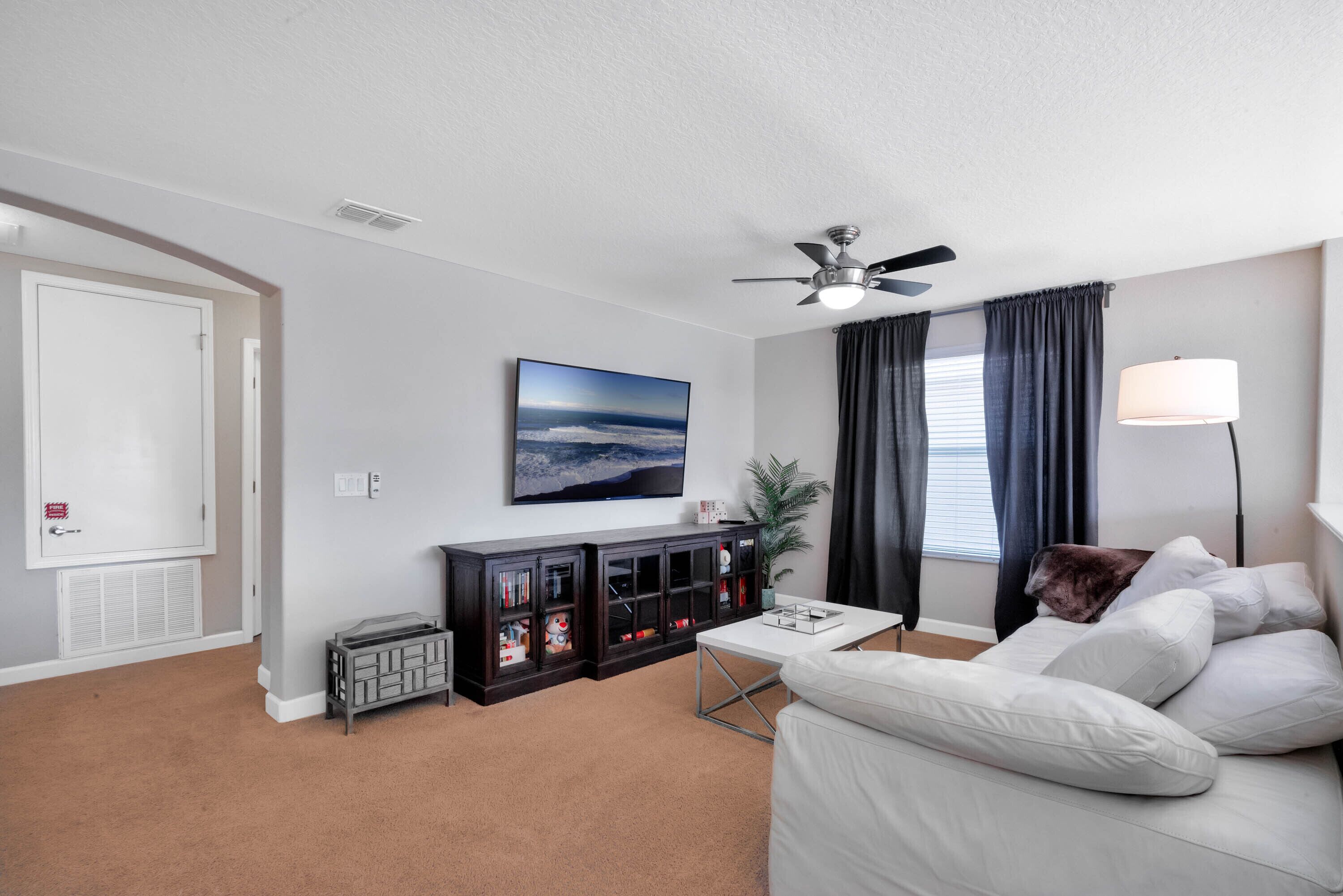 Een woonkamer met een plafondventilator, een flatscreen-tv, een donkere entertainmentkast, een witte bank en een lamp.