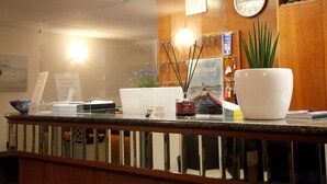 A reception area with a wooden counter, a clock, a potted plant, and a poster on the wall. - Hotel Alfa (Rimini)