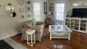 A living room with a beige sofa, a glass coffee table, a white side table, and a television on a cabinet. - Beautiful old style beach cottage 
Walk to beach/ restaurants 
Center of town (Branford)