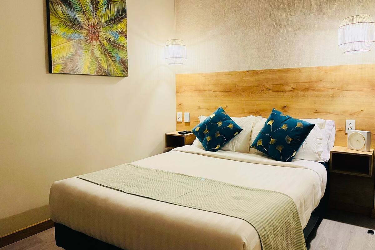 Une chambre d’hôtel comprenant un lit, deux tables de chevet, une petite table, une lampe fixée au mur et un tableau.