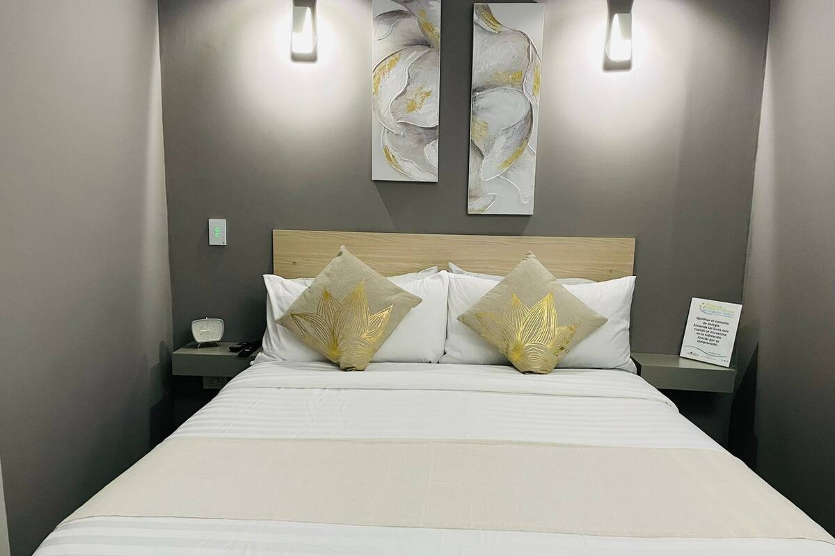 Un lit bien fait, agrémenté de deux coussins décoratifs, une table de chevet avec une horloge et deux lampes fixées au mur.