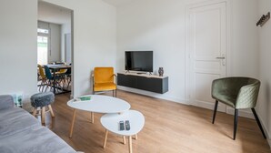A modern living room with a sofa, two white tables, a yellow chair, a green armchair, a TV on a black cabinet, and a dining area in the background. - A bright cocoon for a pleasant stay in Arcachon (Arcachon)