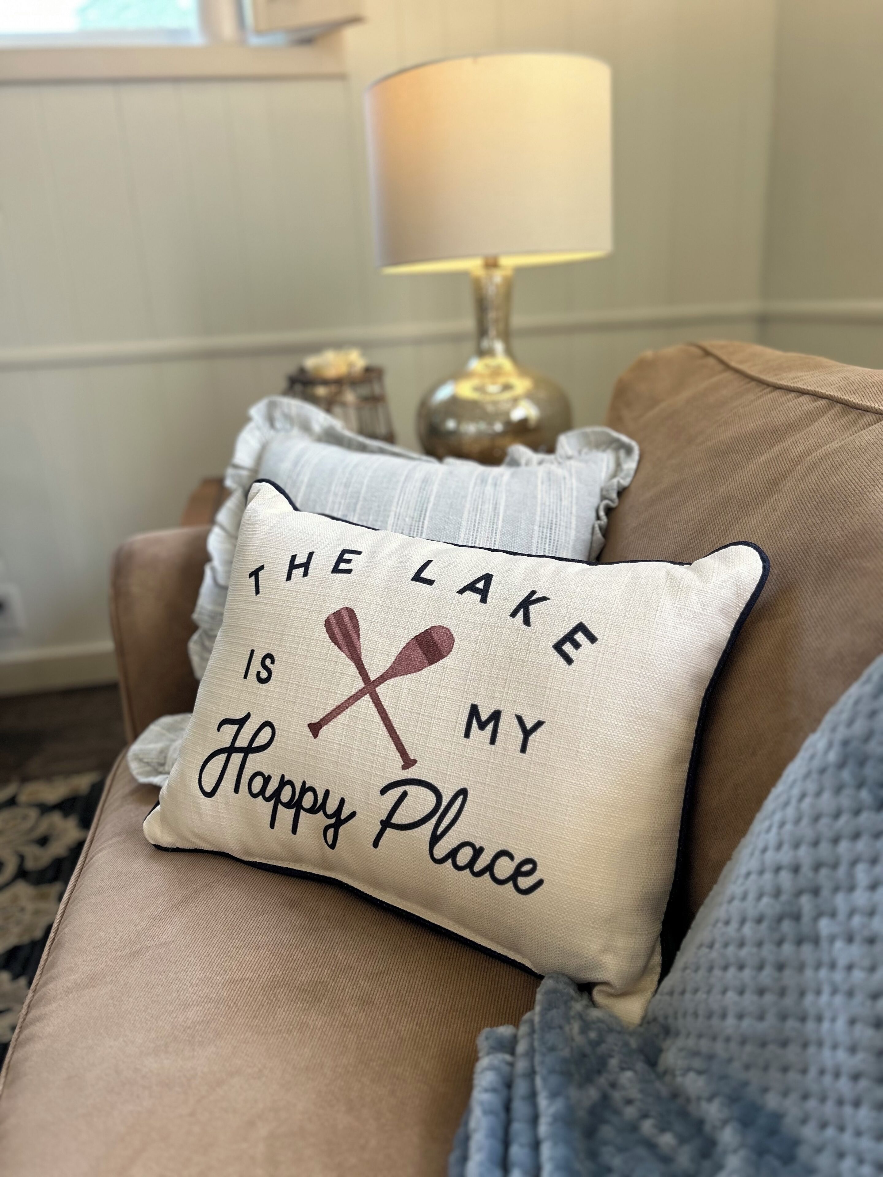 Un coussin beige orné du texte « LE LAC EST MON HAVRE DE PAIX » et de deux pagaies croisées, posé sur un canapé marron.