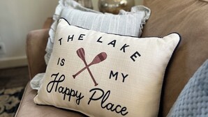 Almofada bege com a frase 'THE LAKE IS MY HAPPY PLACE' e duas pás cruzadas, colocada em um sofá marrom.