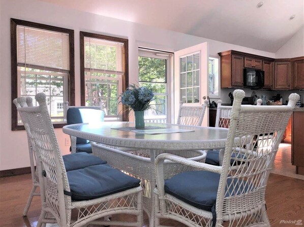A dining area with a round table and wicker chairs, a vase with flowers, and a view of the outdoors through large windows.