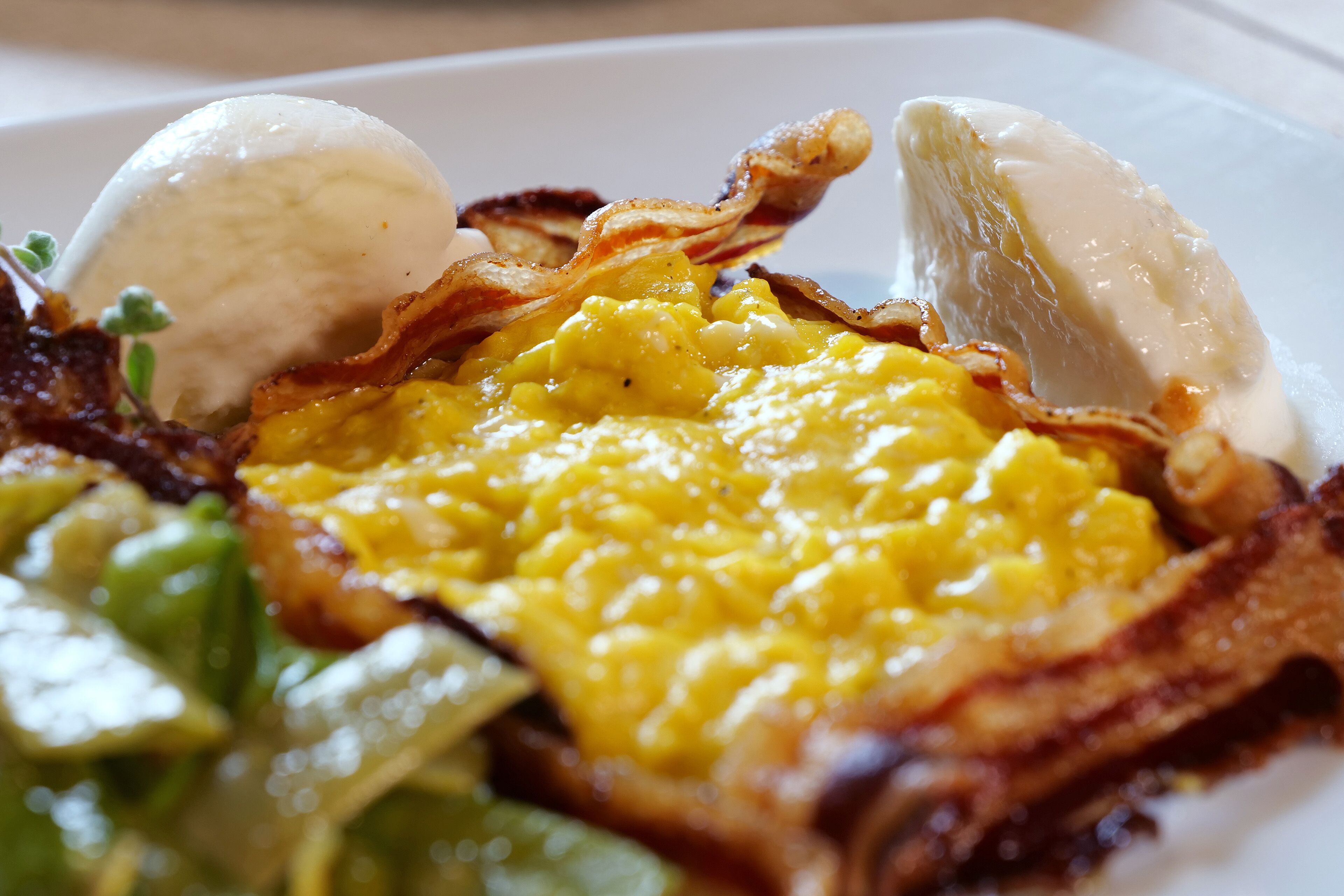 A close-up of a breakfast dish with scrambled eggs, bacon, and a poached egg on a white plate.