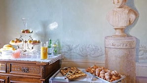Una mesa de buffet con pasteles, frutas y bebidas, una escultura tipo busto identificada como 'DANTE', y un mueble de madera con cajones.