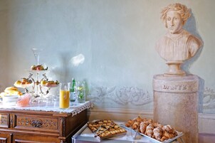 Una mesa de buffet con pasteles, frutas y bebidas, una escultura tipo busto identificada como 'DANTE', y un mueble de madera con cajones.