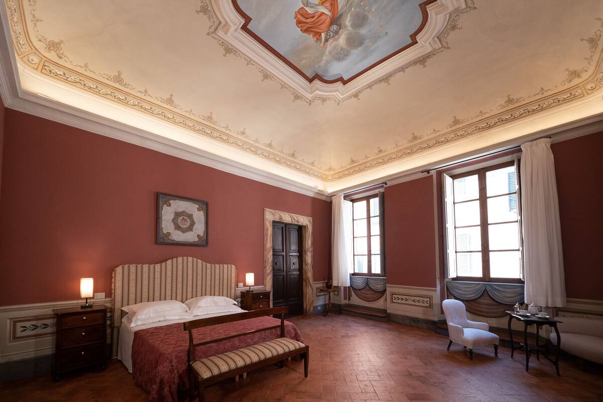Une chambre avec un grand lit, une chaise, une petite table et une fenêtre avec des rideaux.