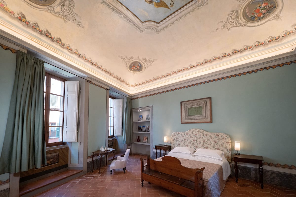Une chambre avec un lit en bois, une chaise blanche, une petite table, une étagère avec des photos et une fenêtre avec des rideaux.