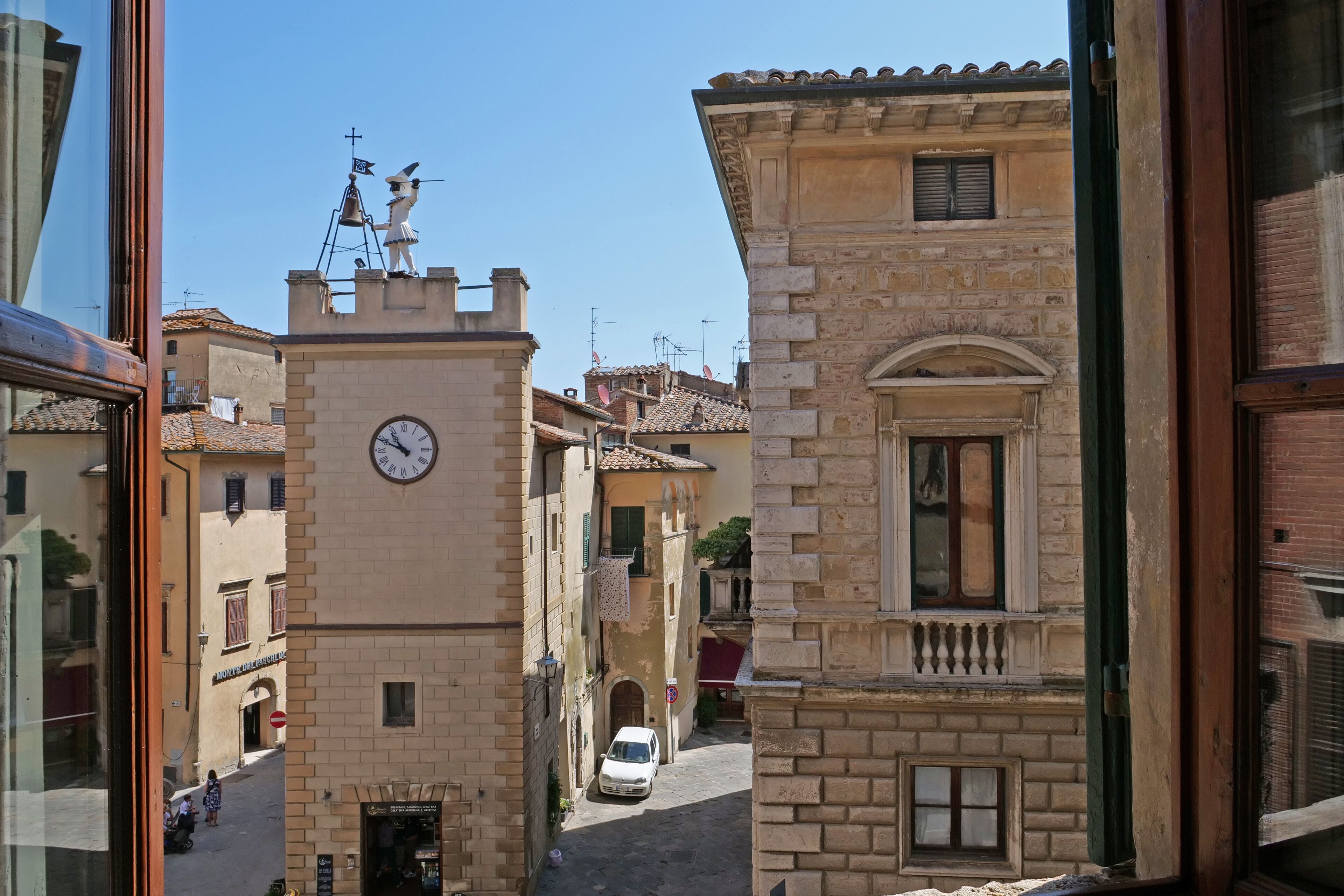 A narrow street with historic buildings, a clock tower, and a white car parked on the side.