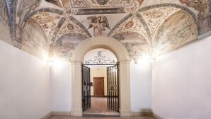 Un pasillo con un techo ornamentado que presenta frescos clásicos, una puerta de madera al final y portones de metal negro.