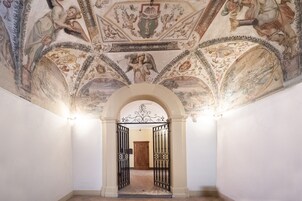 A hallway with an ornate ceiling featuring classical frescoes, a wooden door at the end, and black metal gates.