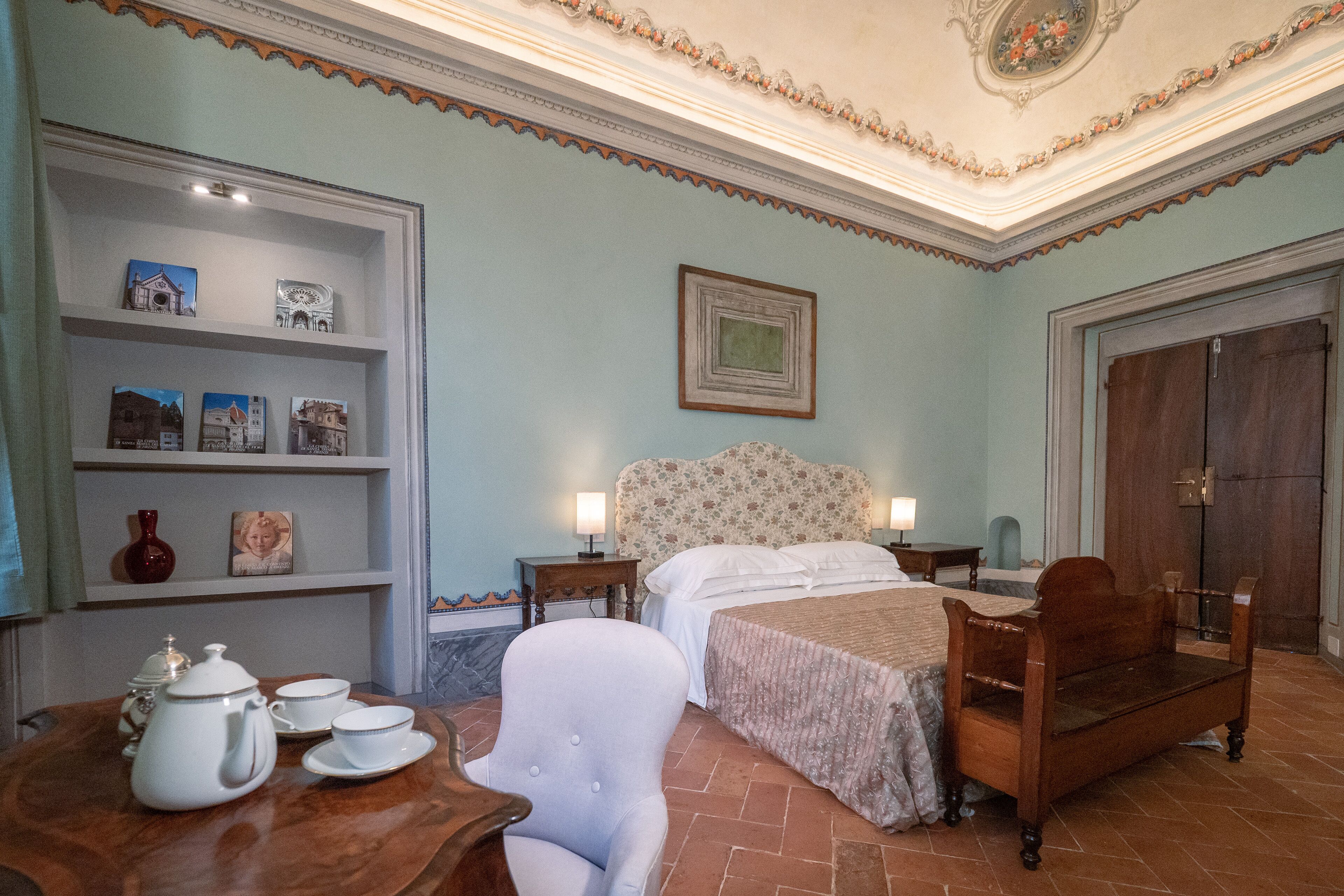 Un dormitorio con una cama, una mesita de noche, una silla, una mesa con tetera y tazas, y un estante con marcos de fotos.