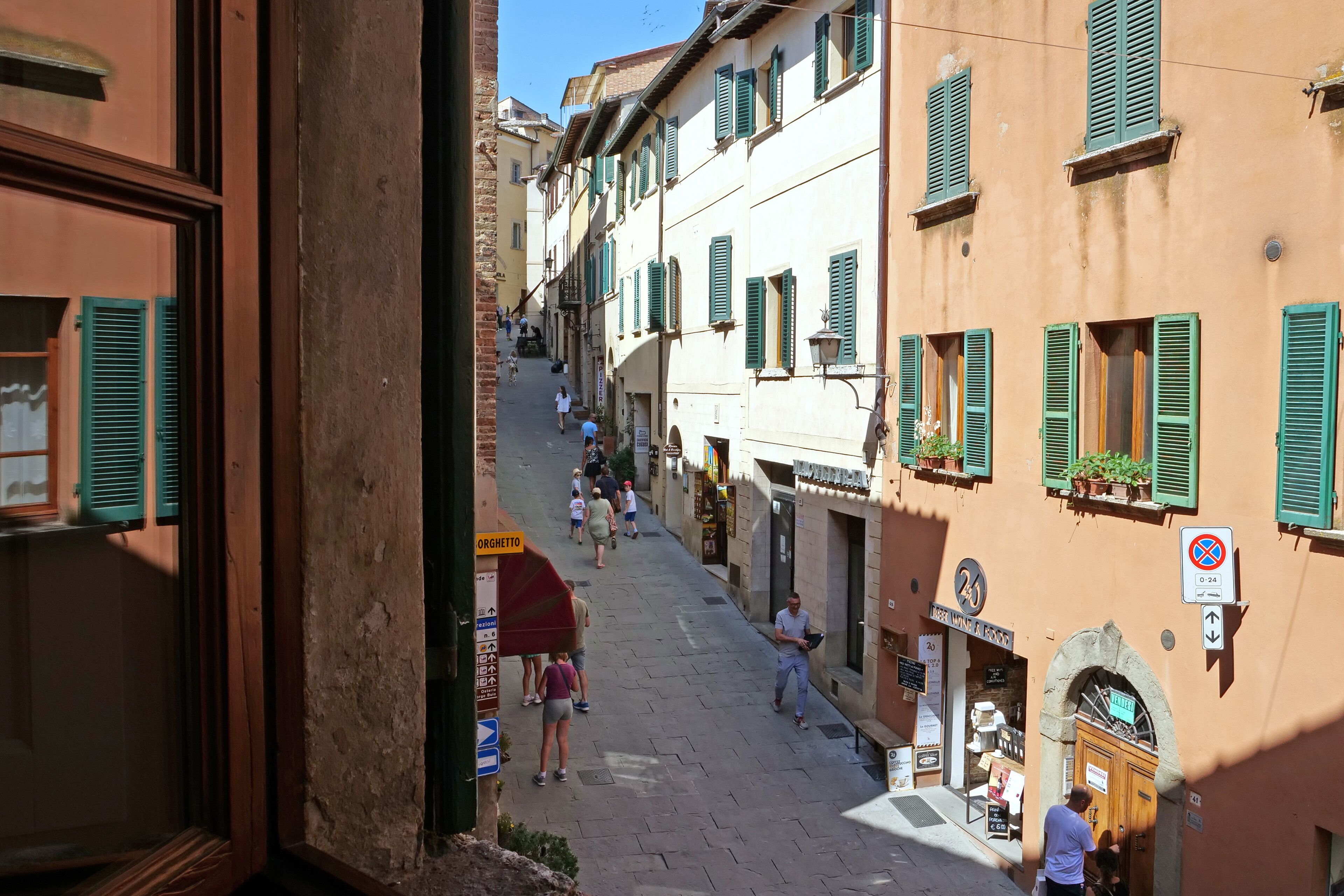 A narrow street with buildings featuring green shutters, a sign for 'Orgetto', and a no parking sign.