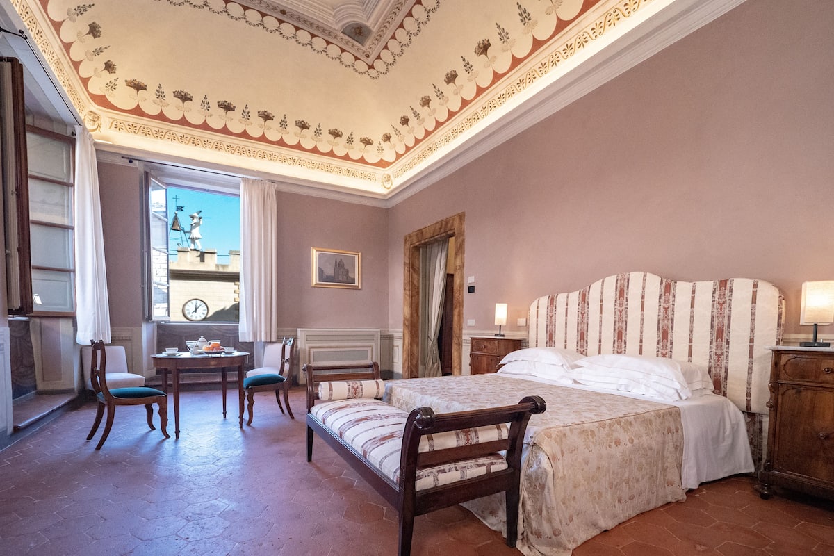 Une chambre à coucher avec un grand lit, un bureau, deux chaises et une fenêtre donnant sur un clocher.