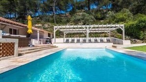 Una piscina rectangular con agua azul cristalina, rodeada por una pérgola blanca con sillones de descanso.
