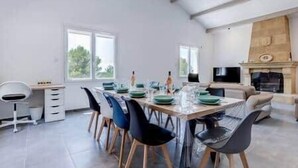 Un comedor moderno con una larga mesa dispuesta para cuatro personas, rodeada de sillas. Se observa un sofá y un televisor al fondo.