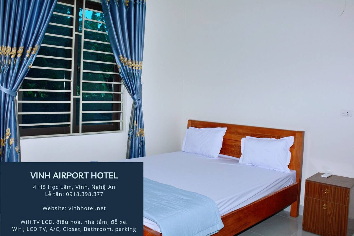 A hotel room with a bed, curtains, a window with blinds, a wooden nightstand, and a small cabinet.