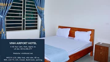 A hotel room with a bed, curtains, a window with blinds, a wooden nightstand, and a small cabinet.