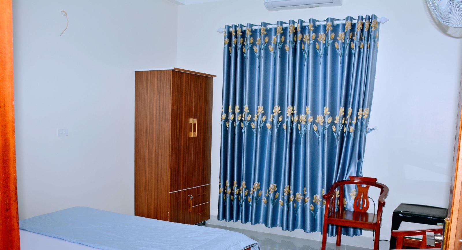 Quarto de hotel com cama, guarda-roupa de madeira, cortina azul com motivos florais, uma cadeira vermelha e escrivaninha com impressora preta.