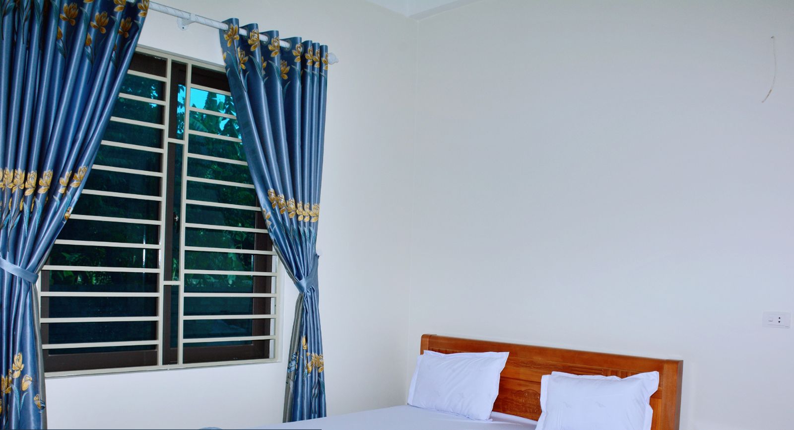 Habitación de hotel con cama, cortinas, ventana con persianas, mesita de noche de madera y un armario pequeño.
