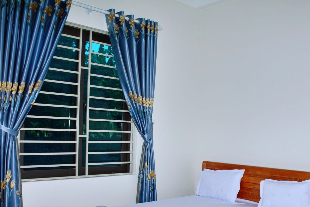 Een hotelkamer met een bed, gordijnen, een raam met zonwering, een houten nachtkastje en een klein kastje.