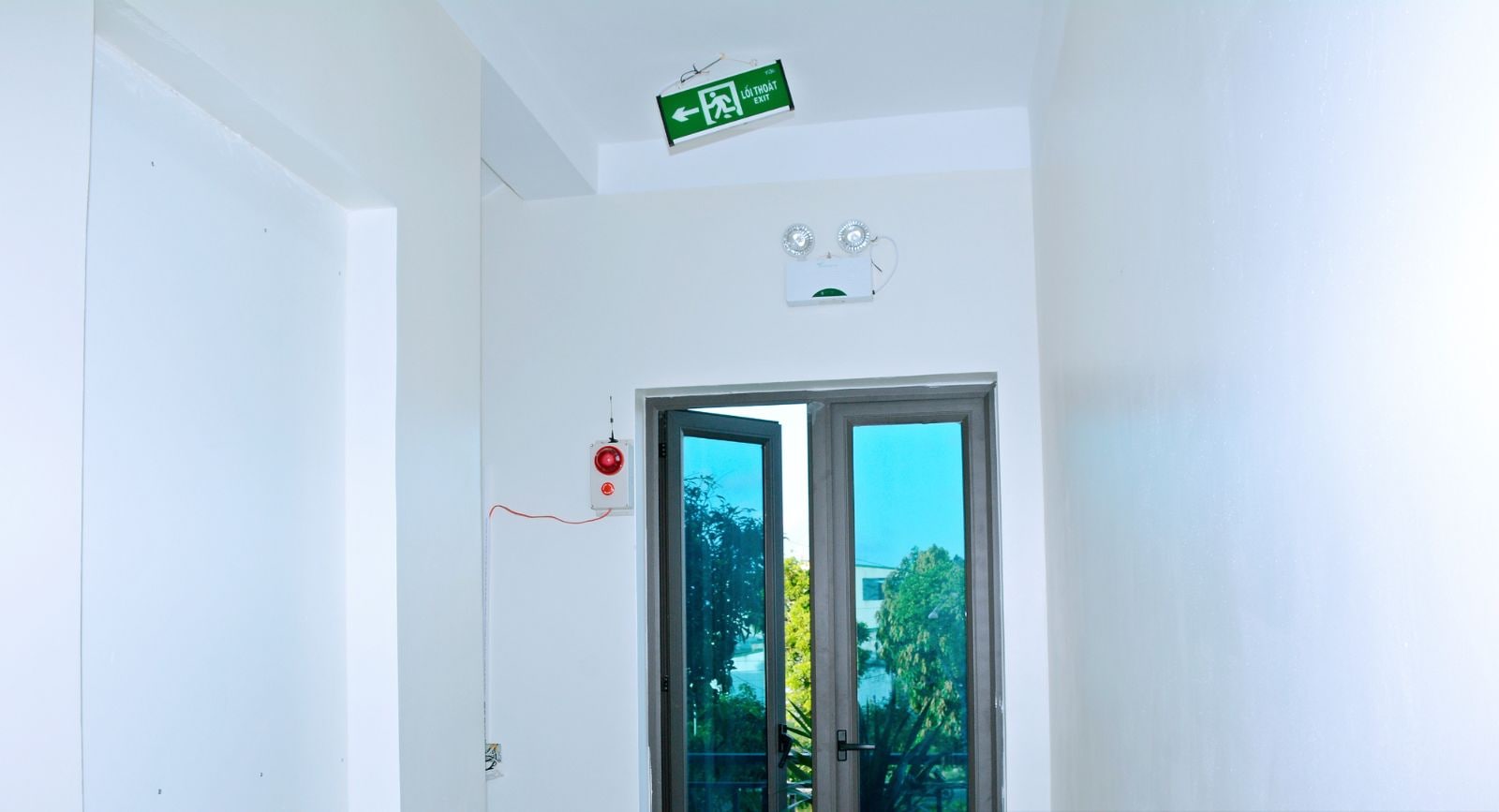 Entrada de un hotel con un letrero de salida de emergencia verde, un botón de emergencia rojo y vista al exterior a través de una puerta de vidrio.