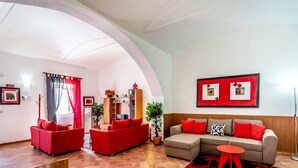 Una sala de estar con un sofá rojo, un sofá gris y una mesita de centro roja. Se observa un mueble de madera, una lámpara de pie y una planta en maceta.