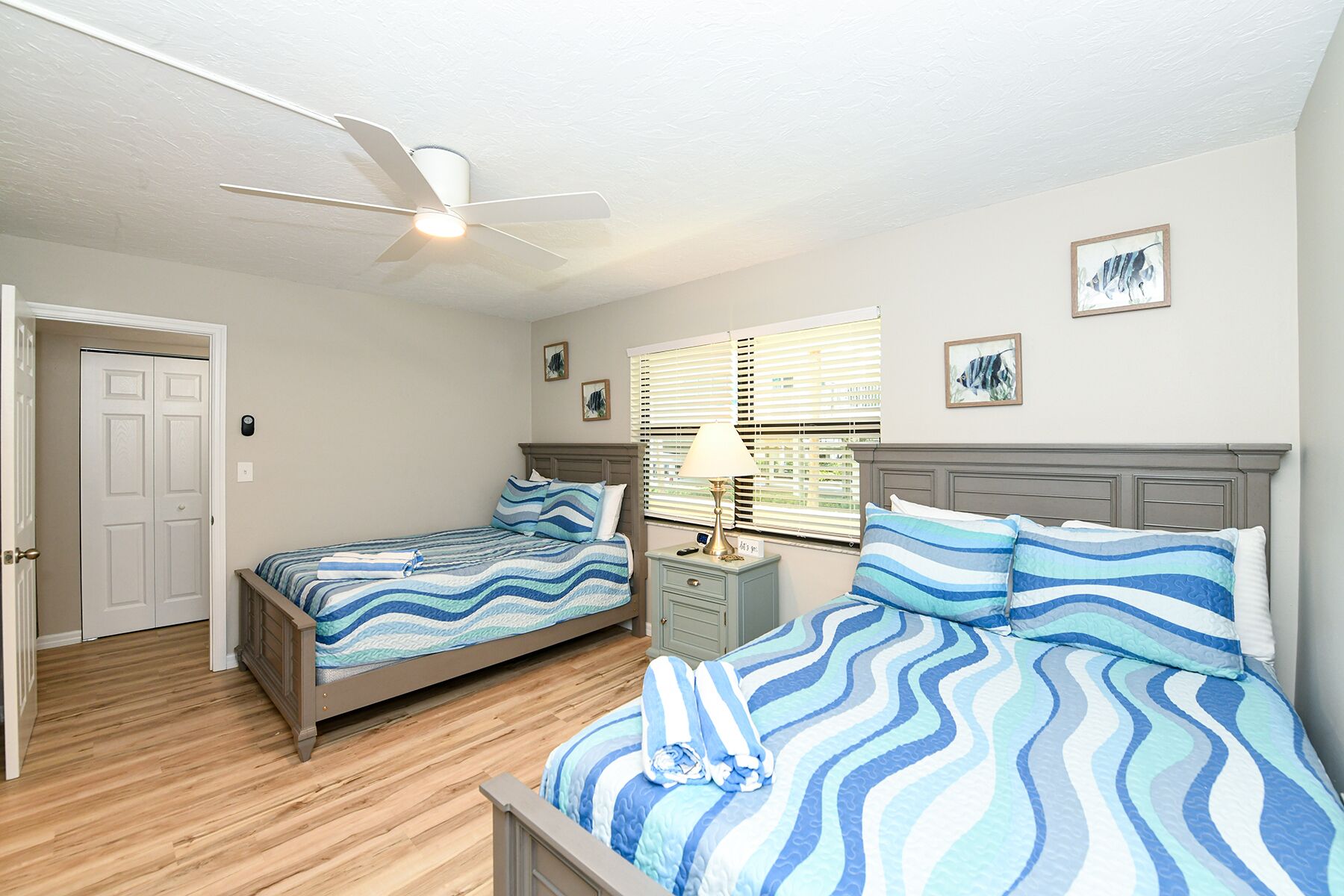 A bedroom with two beds, a ceiling fan, a nightstand with a lamp, and a window with blinds.