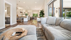 A spacious living room with a large grey sofa, a wooden coffee table with remotes, and a view of the kitchen and dining area in the background. - Luxury LAKEFRONT home  Insane view POPULAR spot 
Sleeps 16+ SERENITY (Kingsland)