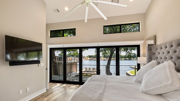 A bedroom with a large bed, a flat-screen TV, and a view of a lake through a sliding glass door. - Luxury LAKEFRONT home  Insane view POPULAR spot 
Sleeps 16+ SERENITY (Kingsland)