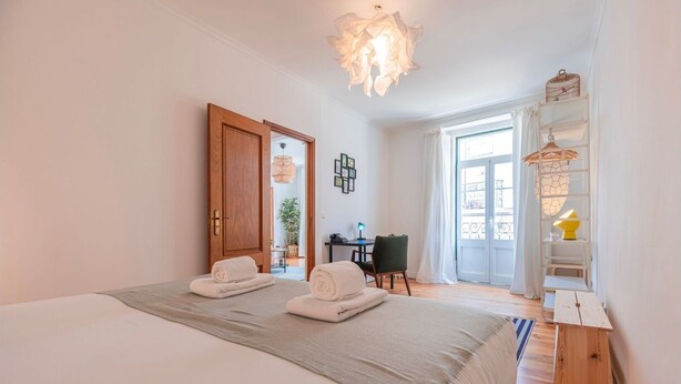Ein Schlafzimmer mit einem großen Bett, zwei zusammengelegten Handtüchern, einer Holztür, einem Schreibtisch mit Stuhl, einem kleinen Tisch mit Lampe und einem Balkon mit Geländer.