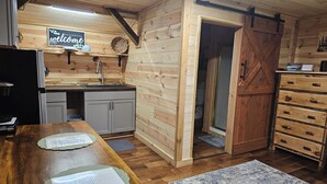 Puinen mökki, jonka sisätiloissa on keittiö, ruokapöytä ruokalistoineen sekä vetolaatikoilla varustettu puinen kaappi.