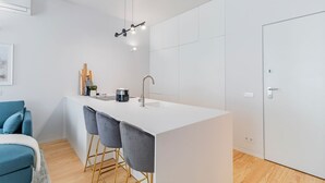 A modern kitchen with a white countertop, a sink, and a coffee machine. There are three grey bar stools and a blue sofa in the adjacent living area. - Almada 551 - 1.1 l Stylish Getaway by LovelyStay (Porto)