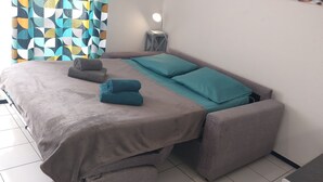 Un lit bien fait, recouvert d’une housse grise, avec deux coussins turquoise et deux serviettes enroulées.