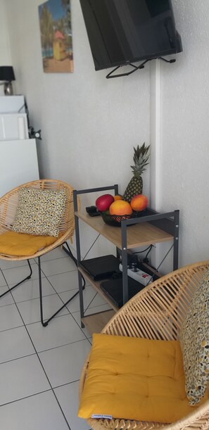Eine Raumecke mit zwei Korbstühlen, einer mit gelber Sitzkissen, einem kleinen Tisch mit Früchten und einem an der Wand montierten Fernseher.
