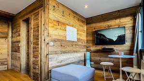Ein gemütliches Zimmer mit Holzwänden, einem wandmontierten Flachbildfernseher, einem kleinen runden Tisch mit zwei Hockern und einer blauen gepolsterten Bank.